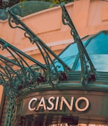 The image displays the entrance to a casino, characterized by detailed and ornate architectural elements with green metal structures and a sign that reads 'CASINO'. The background includes a glass dome and a peach-colored wall.