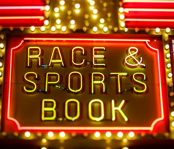 Bright neon lights on a marquee sign display the words 'RACE & SPORTS BOOK'. The sign is surrounded by rows of yellow and white lights against a red background, evoking a vibrant, energetic atmosphere typically associated with casinos or entertainment venues.