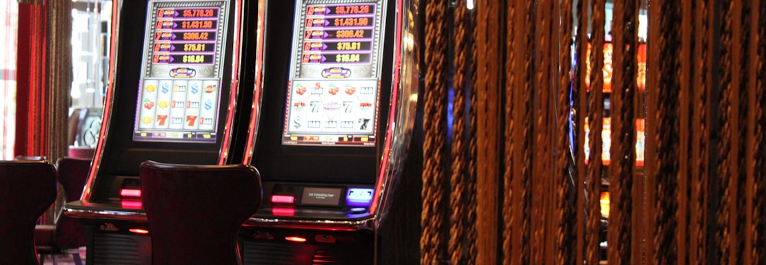 A pair of casino slot machines, each with detailed screens displaying various game outcomes and jackpot amounts. The chairs in front of the slot machines are dark and cushioned for comfort. The background features a hanging curtain made of golden ropes and other casino decorations are visible.