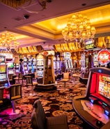 A vibrant casino interior filled with rows of colorful slot machines, each illuminated with bright lights and displays. The ceiling is adorned with elegant chandeliers and golden drapes, creating a luxurious atmosphere. The patterned carpet adds to the elaborate design of the space.