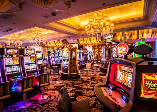 A vibrant casino interior filled with rows of colorful slot machines, each illuminated with bright lights and displays. The ceiling is adorned with elegant chandeliers and golden drapes, creating a luxurious atmosphere. The patterned carpet adds to the elaborate design of the space.