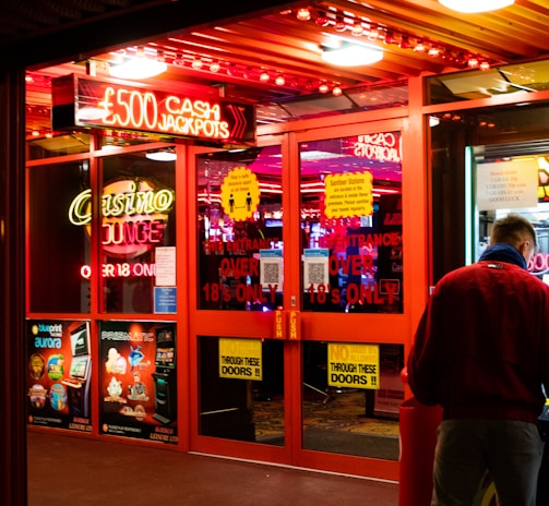 A brightly lit entrance to a casino with neon signs, including one advertising a £500 jackpot. The doors have clear signage indicating entry is restricted to those over 18. A person in a red jacket stands to the right, possibly interacting with a machine or display.