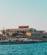 A seaside view of a large building labeled as a casino, situated on a rocky shore. The building is surrounded by palm trees and other smaller structures. The calm sea water reflects the sky, and there are several closed umbrellas lined along the waterfront.