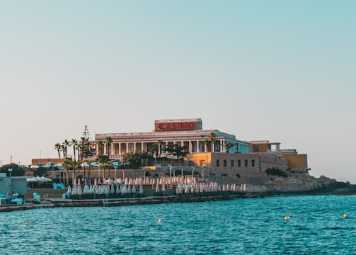 A seaside view of a large building labeled as a casino, situated on a rocky shore. The building is surrounded by palm trees and other smaller structures. The calm sea water reflects the sky, and there are several closed umbrellas lined along the waterfront.