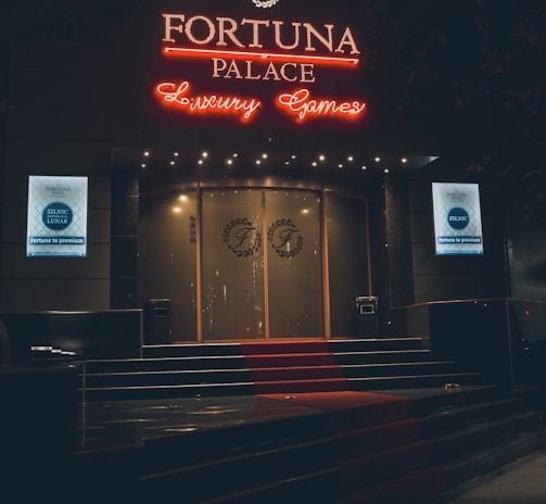 A neon-lit casino entrance with a red carpet leading up to large double doors. The sign reads 'Fortuna Palace Luxury Games' in bright red and white neon lights. The dimly lit atmosphere suggests a night-time setting. There are two digital displays on either side of the entrance showing promotional content.